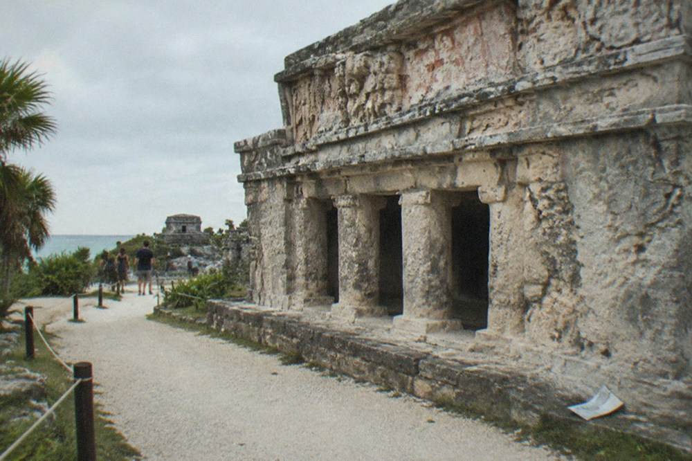 A stone Maya temple building with carved stucco detail beside a walking path