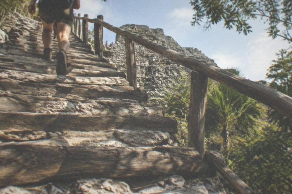 Traveler wearing hiking shoes and a daypack on the wooden staircase of Nohoch Mul pyramid.