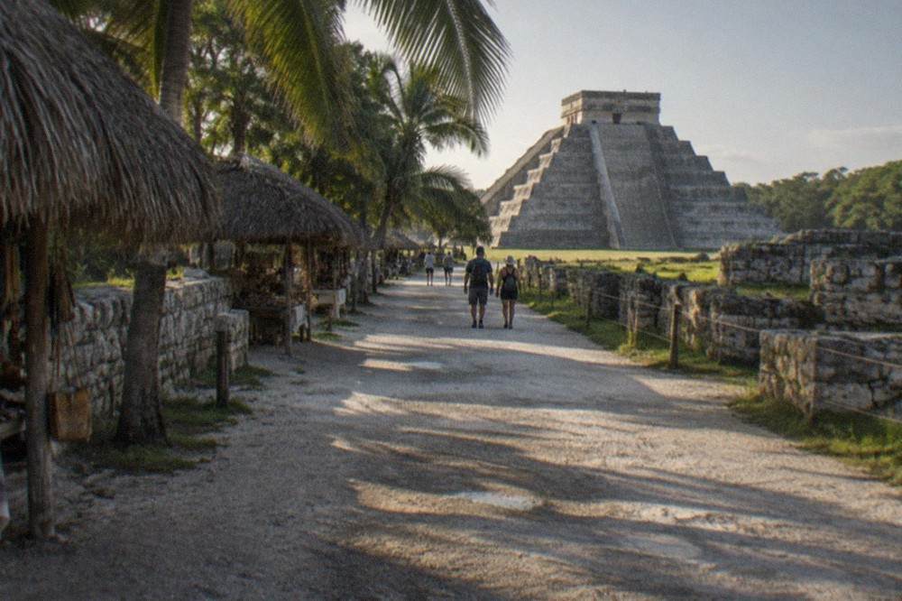 A long white stone walkway lined with low ruins and vendor palapas under palm trees