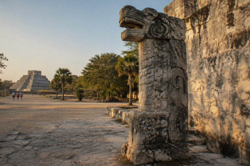 Weathered stone serpent column carved with a feathered serpent standing beside a ruined wall