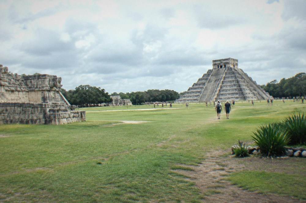 A wide grassy plaza with stone ruins and a stepped pyramid under scattered clouds
