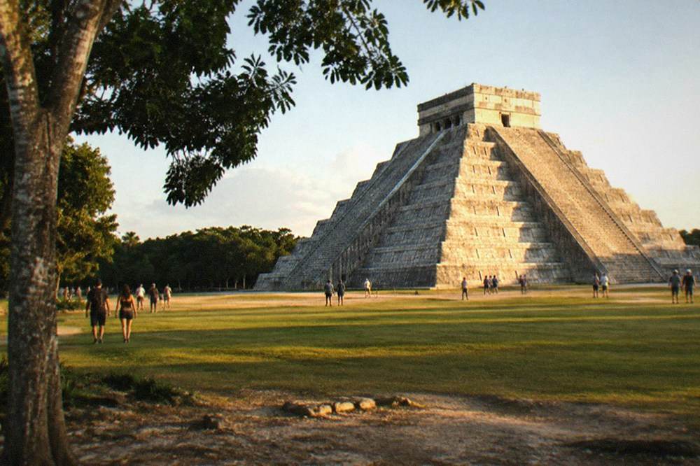 A tall stepped stone pyramid rising above a grassy plaza ringed by trees