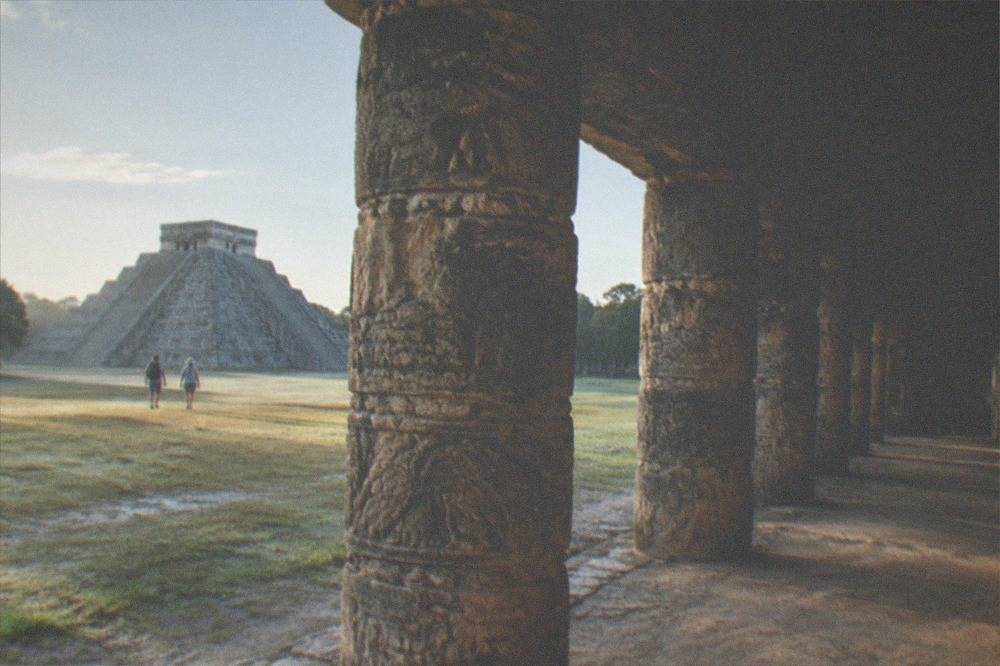 Visitors walking a wide stone plaza beside a carved Maya column complex in morning light