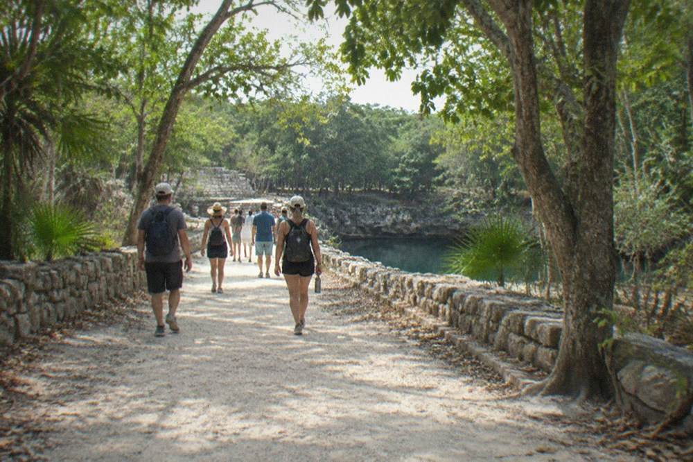 A small tour group walking a dirt path between stone ruins under tall trees at Chichen Itza