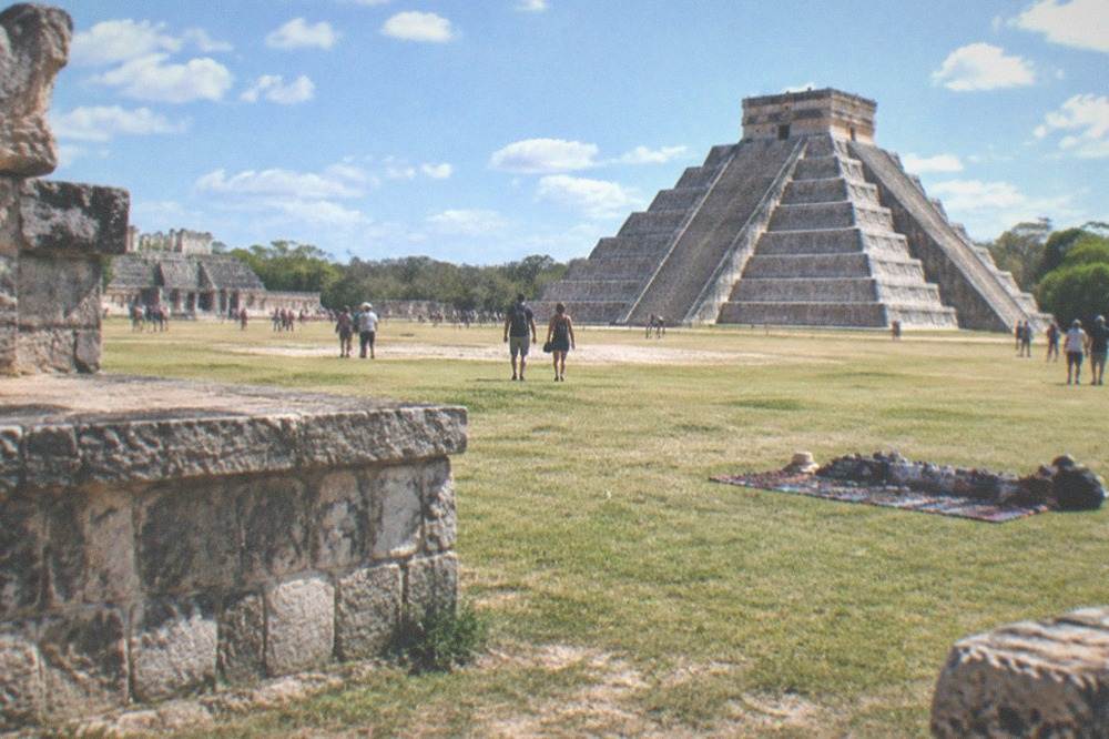 A wide plaza of Mayan stone buildings under a bright morning sky