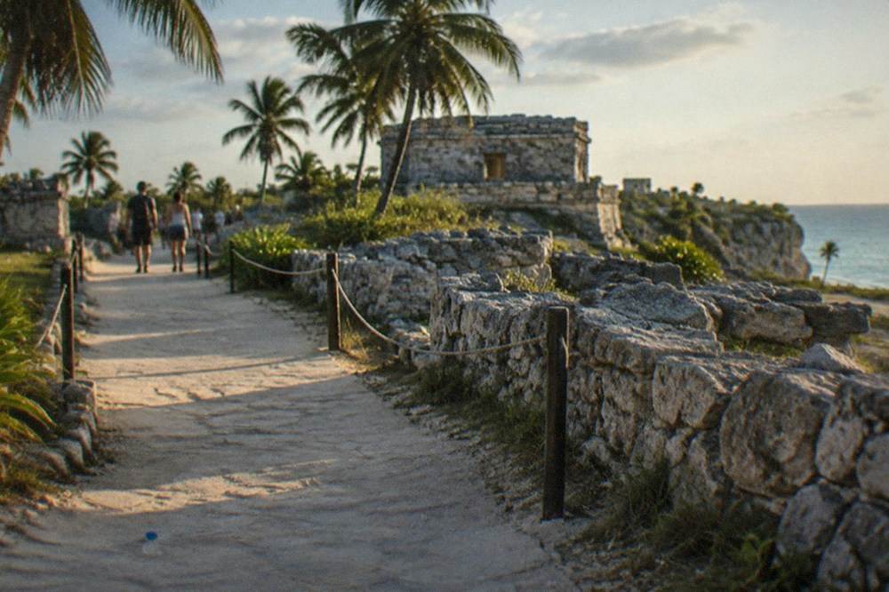 Visitors walking a paved path between low stone ruins walls under palm trees