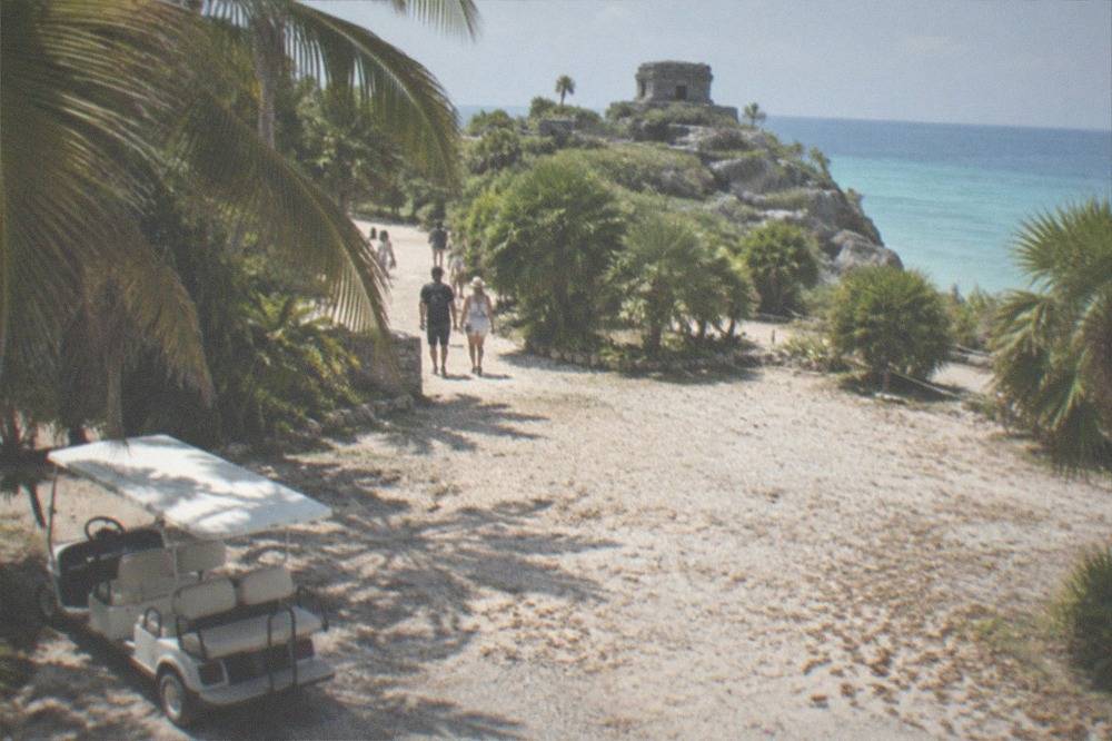 Palm-lined gravel parking area with a shuttle cart near the Tulum ruins entrance